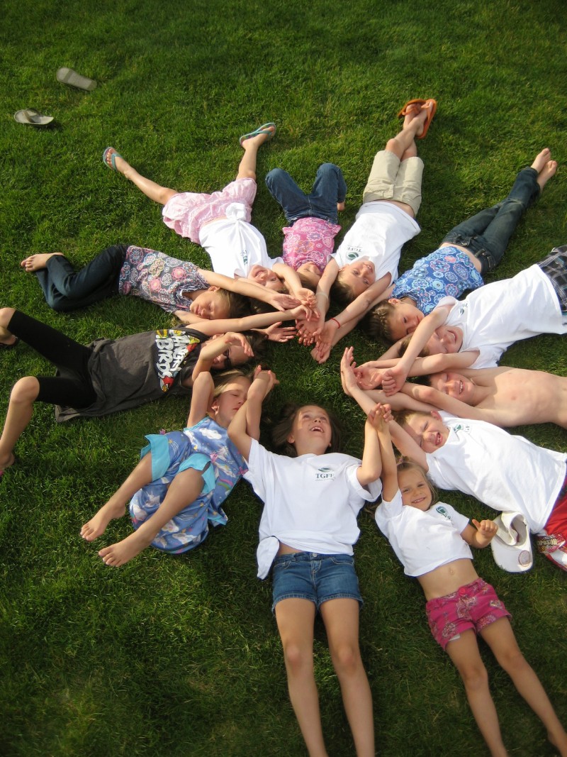 A group of young TGFF nieces and nephews lay on the grass in a circle with their hands touching each other.