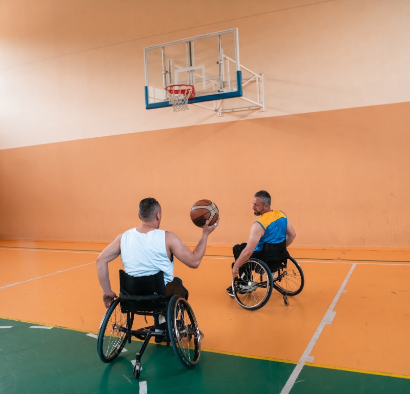 Two gentlemen in wheelchairs playing basketball in a gymnasium.