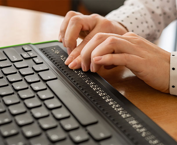 A person uses a computer with a braille display and a computer keyboard.