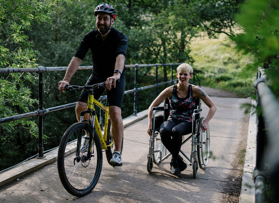 Two people enjoying time outdoors on a boardwalk trail. One person is on a bike and the other is in a wheelchair